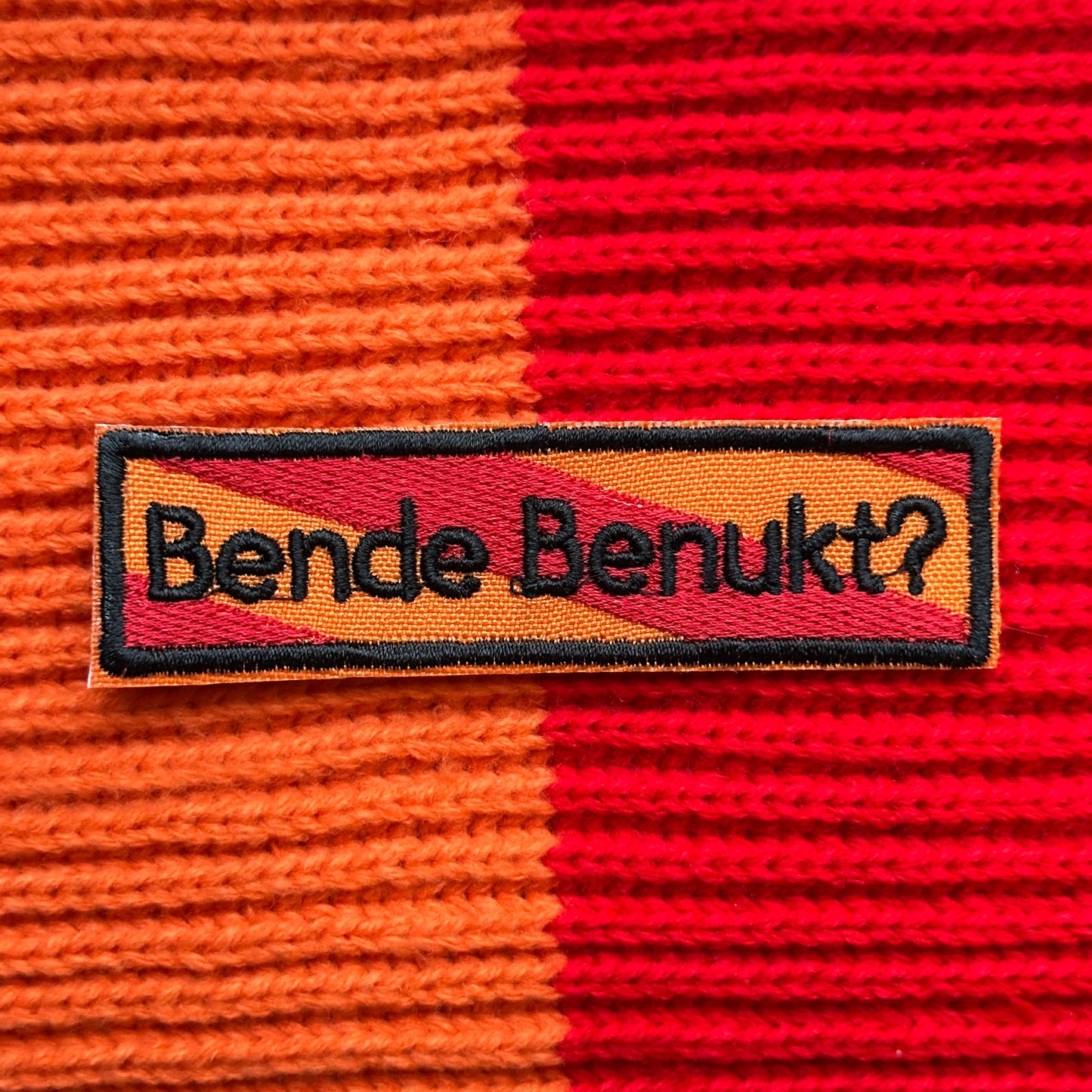 Een rechthoekig spreukenbord, met een rood-oranje gestreepte achtergrond in de Kielegat kleuren. Hierop staat de tekst 'Bende benukt?'.