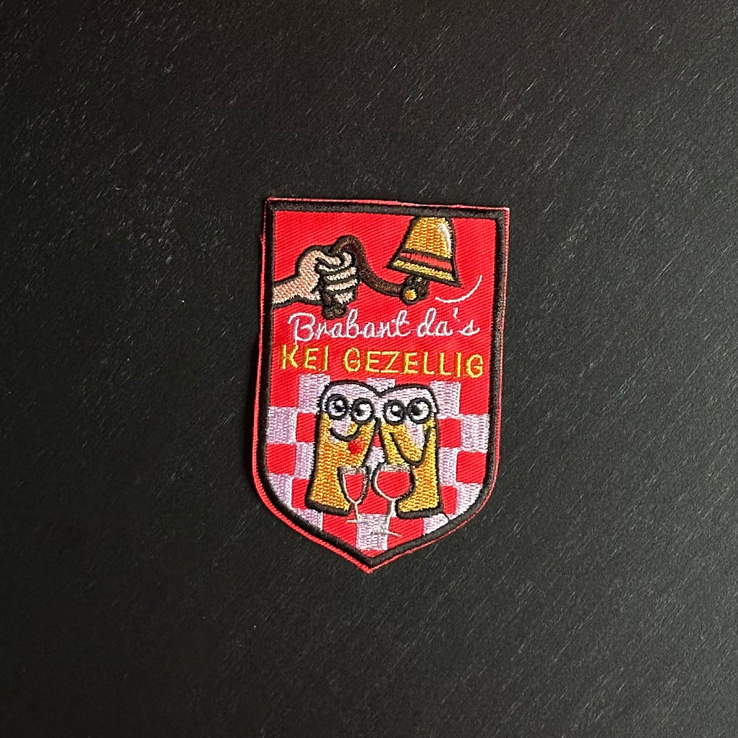 Een rood embleem, waarbij er aan een bel getrokken word, twee biertjes gezellig met een wijntje zitten en de tekst 'Brabant da's KEI GEZELLIG' is opgenomen.