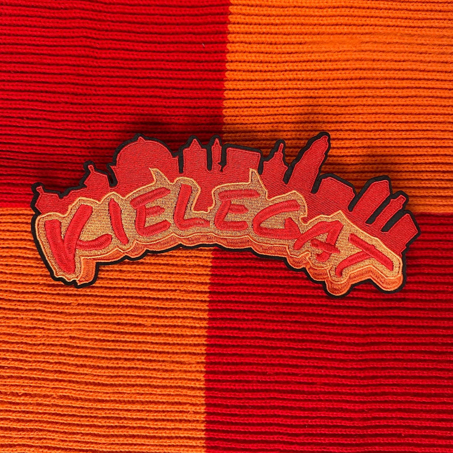 Het Kielegat (rug)embleem. Dit embleem heeft de tekst Kielegat in het rood, met een oranje achtergrond. Daarboven is ook in het rood de kenmerkende Bredase gebouwen opgenomen.