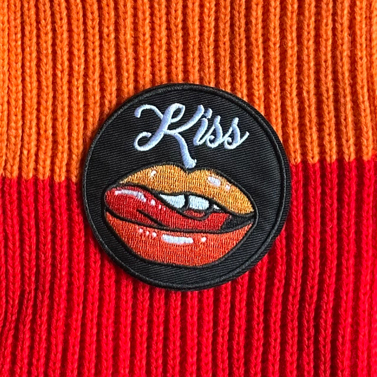 Rond Kielegat embleem met een zwarte achtergrond, en beeld rood-oranje met de tong likkende lippen. Hiernaast is het woord 'Kiss' opschreven.