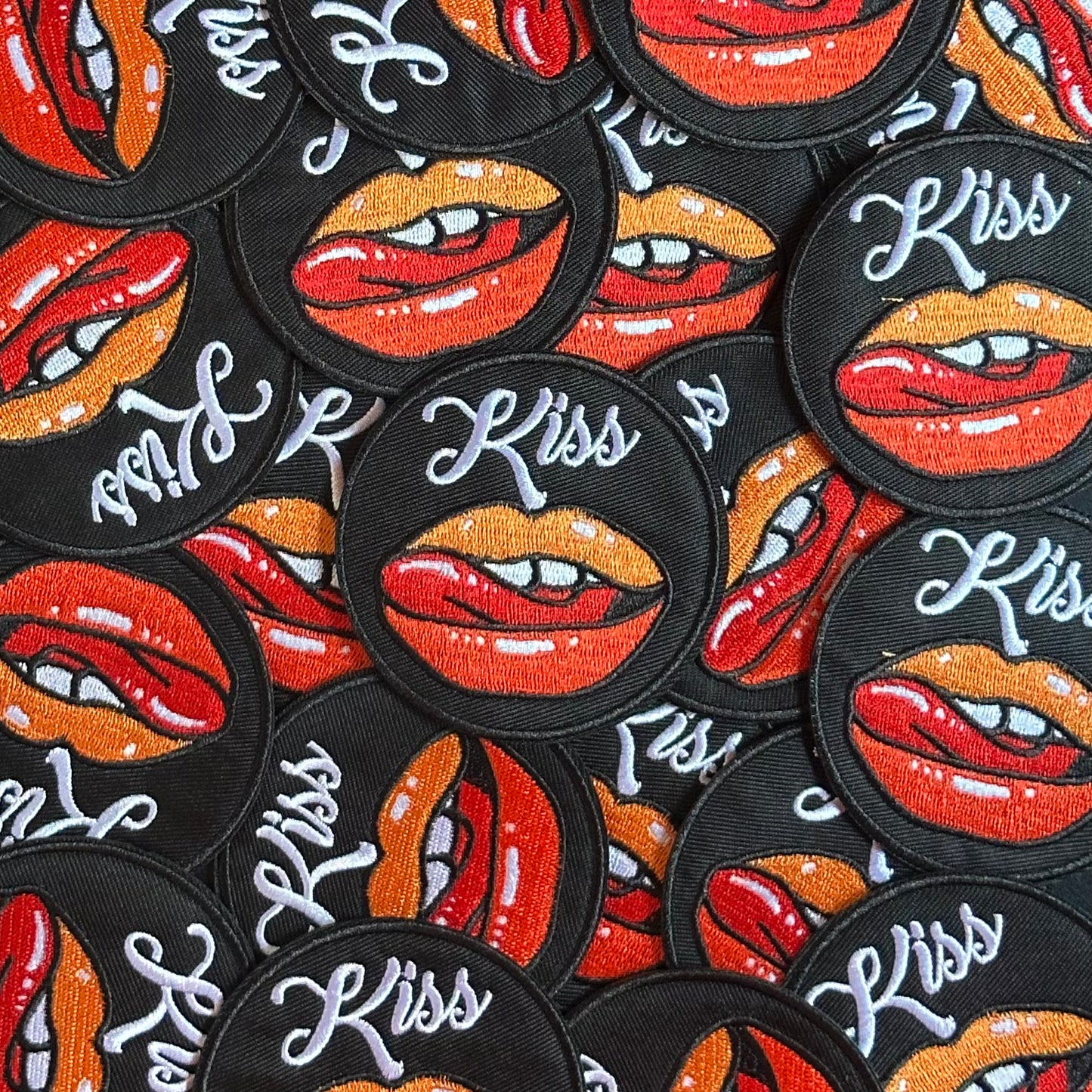 Rond Kielegat embleem met een zwarte achtergrond, en beeld rood-oranje met de tong likkende lippen. Hiernaast is het woord 'Kiss' opschreven.