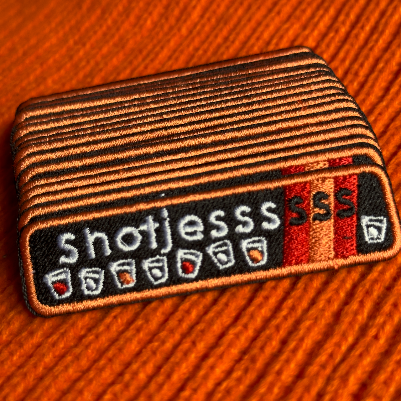 Een rechthoekig Kielegat embleem met een zwarte achtergrond waarop rood en oranje accenten zijn aangebracht en de tekst 'Shotjesssss' is opgenomen en shotglazen zijn afgebeeld.