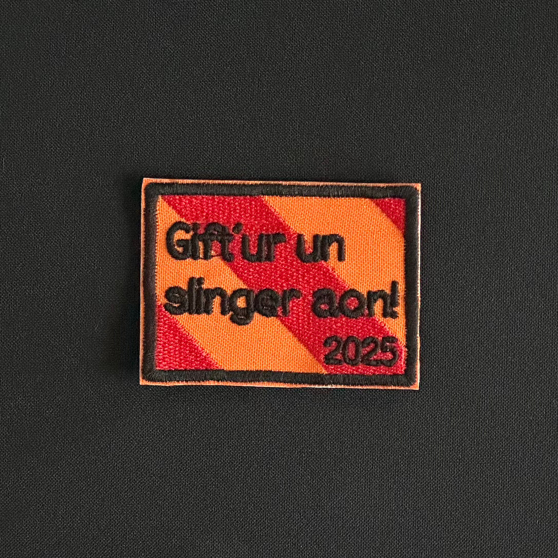 Een rechthoekig spreukenbord, met een rood-oranje gestreepte achtergrond in de Kielegat kleuren. Hierop staat de tekst 'Gift'ur un slinger aon! 2025'.