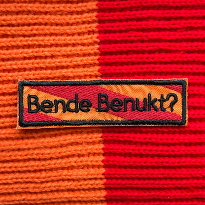 Een rechthoekig spreukenbord, met een rood-oranje gestreepte achtergrond in de Kielegat kleuren. Hierop staat de tekst 'Bende benukt?'.