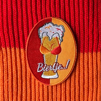 Ovaalvormig Kielegat embleem met rood en oranje achtergrond, waarop een biertje met tieten is afgebeeld, waarbij de tieten een rood - oranje gekleurde BH draagt. Ook is de tekst 'Biertje' op het embleem opgenomen.