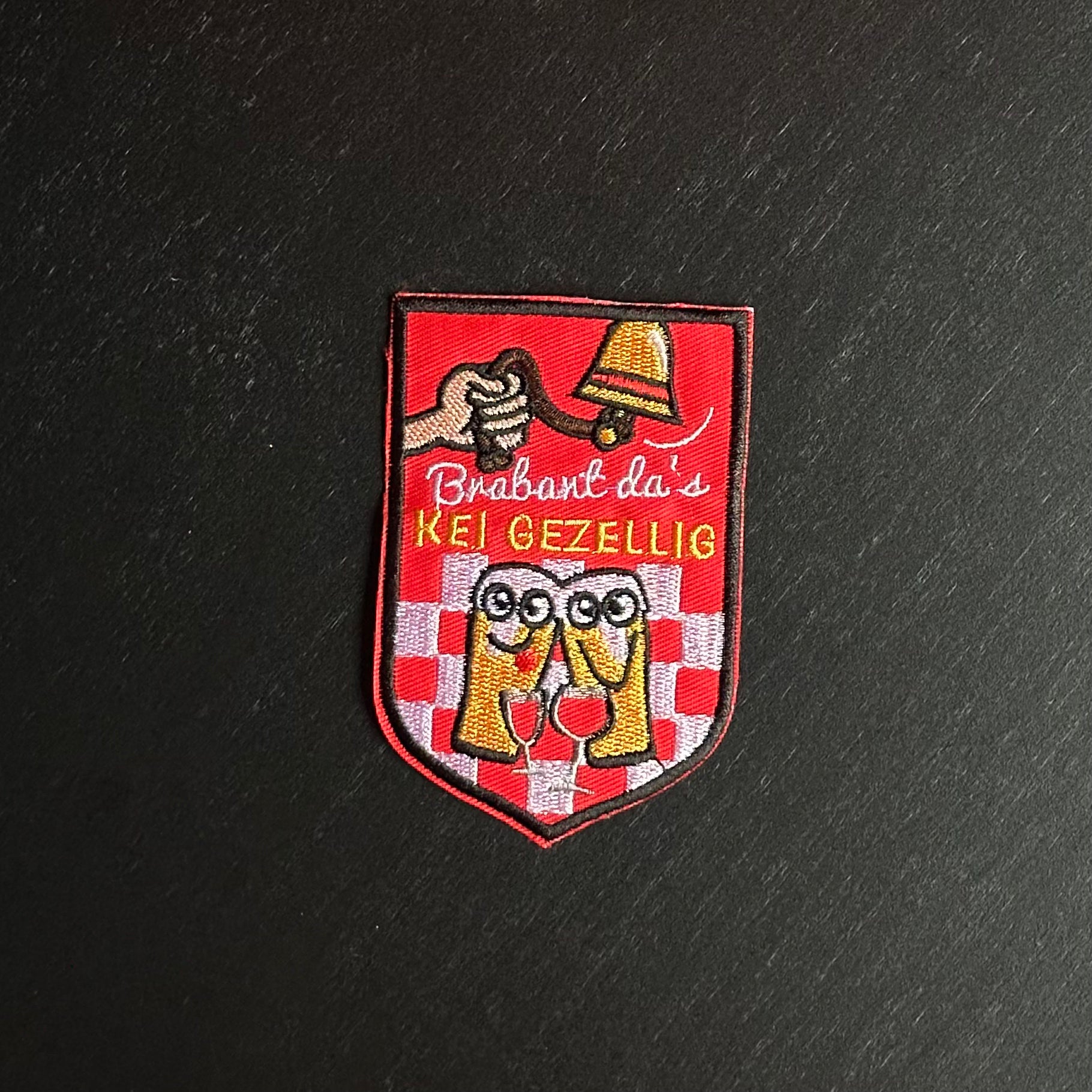 Een rood embleem, waarbij er aan een bel getrokken word, twee biertjes gezellig met een wijntje zitten en de tekst 'Brabant da's KEI GEZELLIG' is opgenomen.