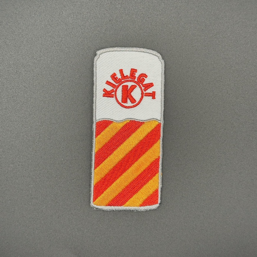 Een blikjes embleem met rood-oranje strepen in de kleuren van het Kielegat, met de tekst ‘Kielegat’ om een K heen gebogen.
