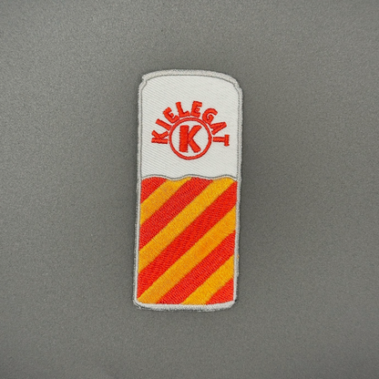 Een blikjes embleem met rood-oranje strepen in de kleuren van het Kielegat, met de tekst ‘Kielegat’ om een K heen gebogen.