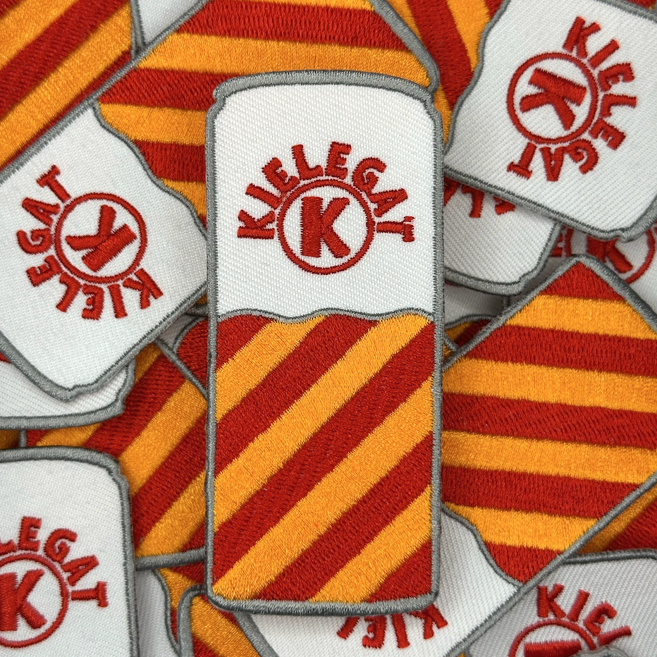 Een blikjes embleem met rood-oranje strepen in de kleuren van het Kielegat, met de tekst ‘Kielegat’ om een K heen gebogen. Groepsfoto