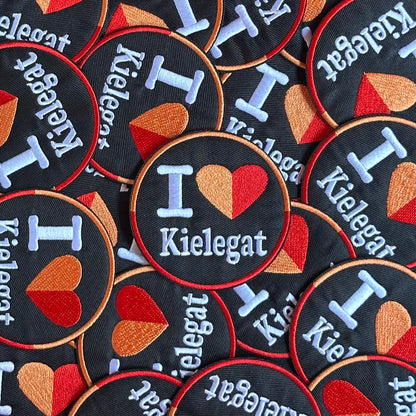 Rond Kielegat embleem met een zwarte achtergrond waar I Love Kielegat op staat. Het hartje is oranje-rood.