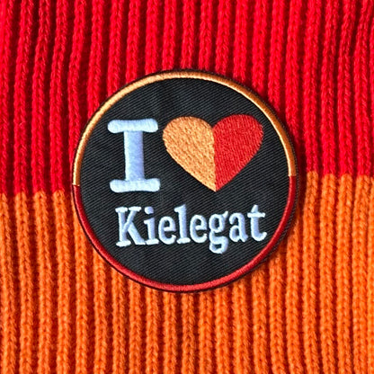 Rond Kielegat embleem met een zwarte achtergrond waar I Love Kielegat op staat. Het hartje is oranje-rood.