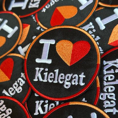 Rond Kielegat embleem met een zwarte achtergrond waar I Love Kielegat op staat. Het hartje is oranje-rood.