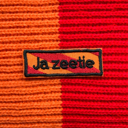 Een rechthoekig spreukenbord, met een rood-oranje gestreepte achtergrond in de Kielegat kleuren. Hierop staat de tekst 'Ja zeetie'.