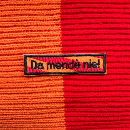 Een rechthoekig spreukenbord, met een rood-oranje gestreepte achtergrond in de Kielegat kleuren. Hierop staat de tekst 'Da mendè nie!'.