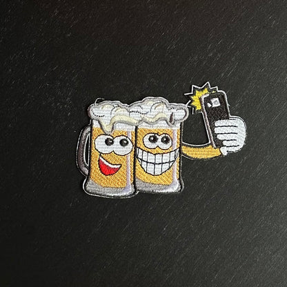 Dit embleem laat twee biertjes zien die samen een vrolijke selfie nemen met een telefoon.