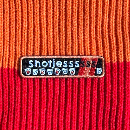 Een rechthoekig Kielegat embleem met een zwarte achtergrond waarop rood en oranje accenten zijn aangebracht en de tekst 'Shotjesssss' is opgenomen en shotglazen zijn afgebeeld.