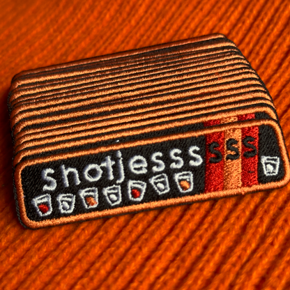 Een rechthoekig Kielegat embleem met een zwarte achtergrond waarop rood en oranje accenten zijn aangebracht en de tekst 'Shotjesssss' is opgenomen en shotglazen zijn afgebeeld.