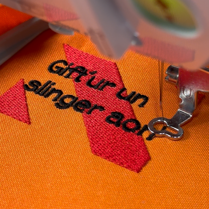 Een rechthoekig spreukenbord, met een rood-oranje gestreepte achtergrond in de Kielegat kleuren. Hierop staat de tekst 'Gift'ur un slinger aon! 2025'.