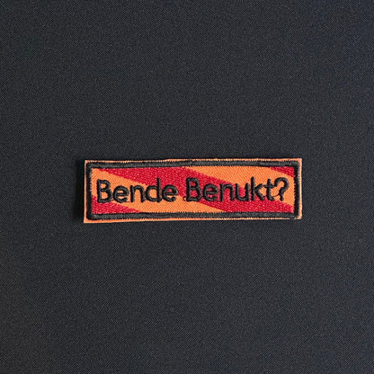 Een rechthoekig spreukenbord, met een rood-oranje gestreepte achtergrond in de Kielegat kleuren. Hierop staat de tekst 'Bende benukt?'.