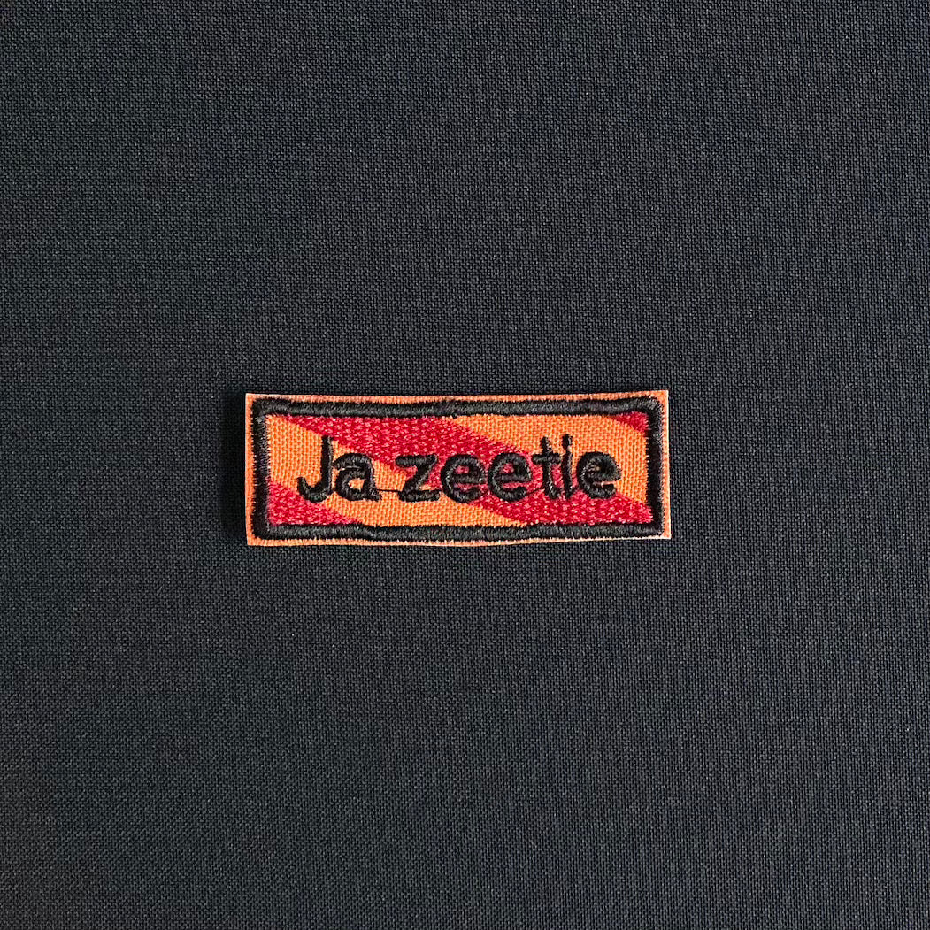 Een rechthoekig spreukenbord, met een rood-oranje gestreepte achtergrond in de Kielegat kleuren. Hierop staat de tekst 'Ja zeetie'.