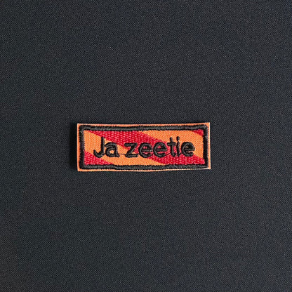 Een rechthoekig spreukenbord, met een rood-oranje gestreepte achtergrond in de Kielegat kleuren. Hierop staat de tekst 'Ja zeetie'.