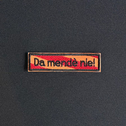 Een rechthoekig spreukenbord, met een rood-oranje gestreepte achtergrond in de Kielegat kleuren. Hierop staat de tekst 'Da mendè nie!'.